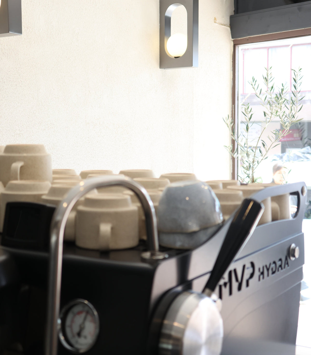 A close-up of a black espresso machine with a silver steam wand and metal portafilter, set in a cafe interior with ceramic cups and a window in the background.